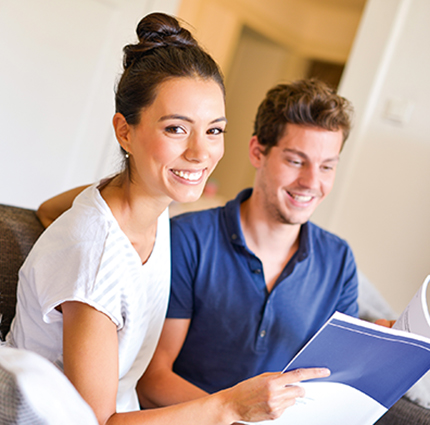 Junge Frau und junger Mann sitzen auf einer Couch und haben ein Studienheft vor sich.