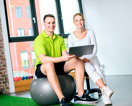 Physiotherapeut und Physiotherapeutin sitzen auf einem Medizinball und halten einen Laptop