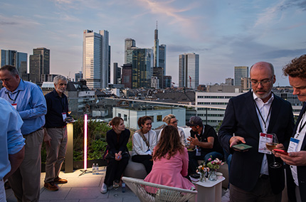 Leute stehen im Abendlicht auf einer Dachterrasse und unterhalten sich.